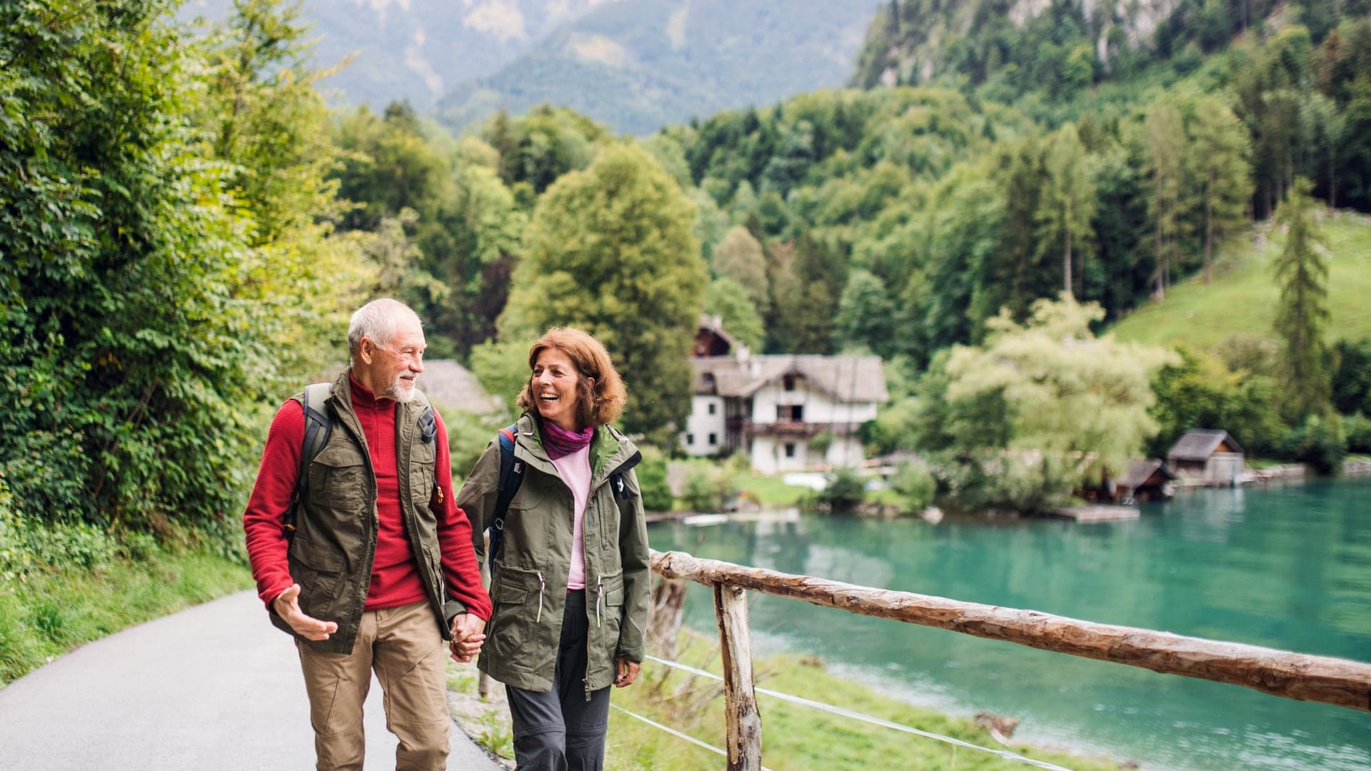 An older couple walking together along a paved path beside a lake, with wooded hills behind them